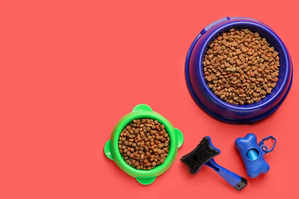 Bowls of dry pet food, grooming brush and waste bags on color background