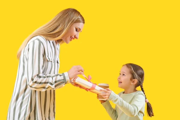 Cute little girl greeting her mother with gift and cup of coffee on yellow background