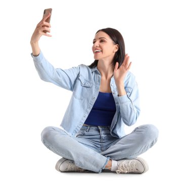 Pretty young woman with smartphone taking selfie against white background