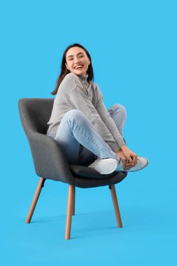 Mavi arka planda siyah koltukta oturan genç bir kadın.