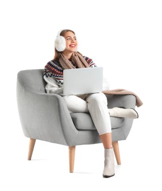 Kulaklıklı genç bir kadın beyaz arka planda gri koltukta dizüstü bilgisayar kullanıyor.