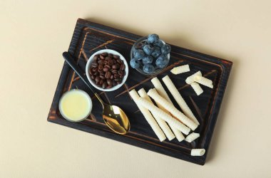 Board with delicious wafer rolls, blueberries, coffee beans and condensed milk on beige background