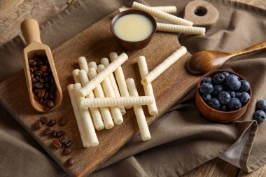 Board with delicious wafer rolls, blueberries and condensed milk on wooden background