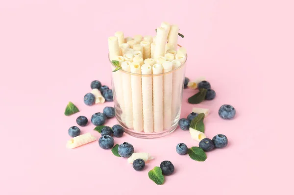Glass with delicious wafer rolls and blueberries on pink background