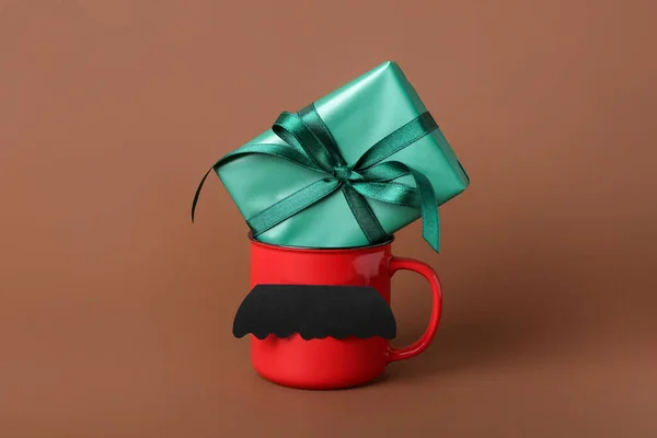 Cup with paper mustache and gift for Father\'s Day celebration on color background