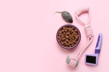 Bir kase ıslak hayvan maması, tımar fırçası ve pembe arka planda oyuncaklar.