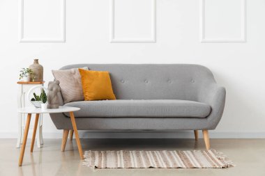 Beyaz duvarın yanındaki masalarda yastıkları ve çiçekleri olan rahat gri kanepe.