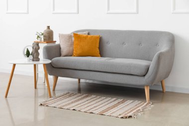 Beyaz duvarın yanındaki masalarda yastıkları ve çiçekleri olan rahat gri kanepe.