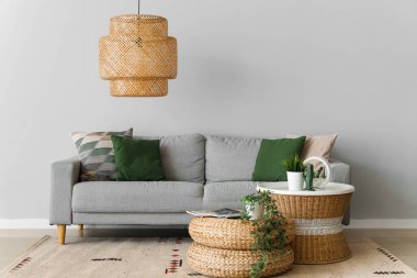 Yastıklı rahat kanepe, ev bitkileri, masa ve hasır pouf, gri duvarın yanında.