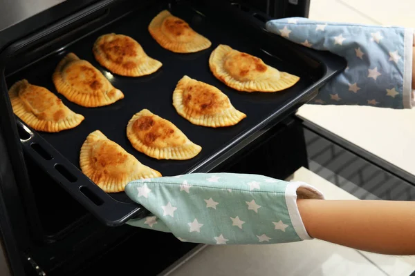 Woman taking baking sheet with tasty meat empanadas out from oven, closeup
