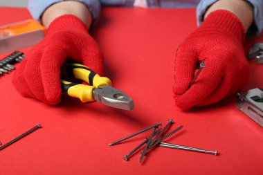 Eldivenli erkek eller kırmızı arka planda pense ve çivi tutuyor.