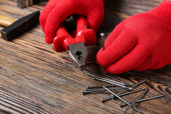 Male hands in gloves holding pliers and nail near wooden table