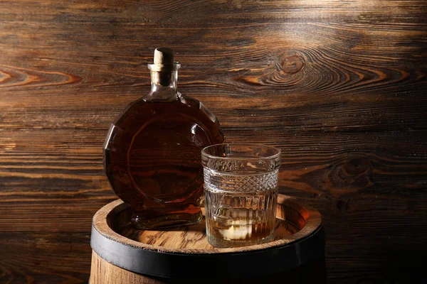 Bottle and glass of cold whiskey on barrel against wooden wooden background