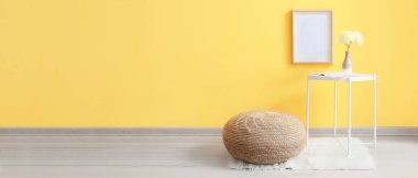 Vazoda içinde çiçekler olan bir masa ve sarı duvarın yanında örülmüş bir puf. Tasarım için pankart