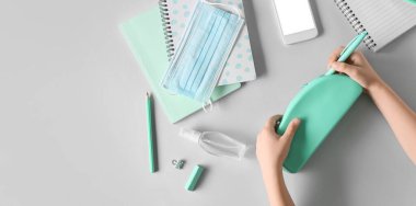 Kalem kutusu, dezenfektan, sağlık maskesi, kırtasiye malzemesi ve cep telefonuyla gri arka planda mesaj için yeri olan çocuk elleri