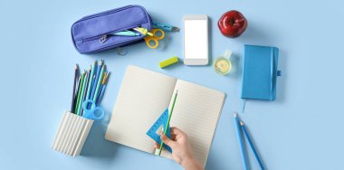 Açık mavi arka planda farklı kırtasiye ve cep telefonuna sahip bir çocuk eli