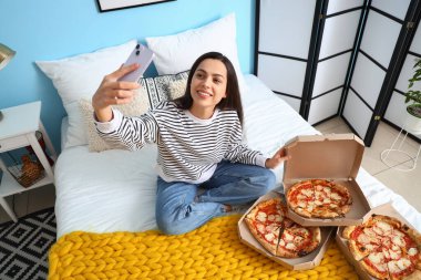 Kutular dolusu lezzetli pizzayla yatak odasında selfie çeken genç bir kadın.