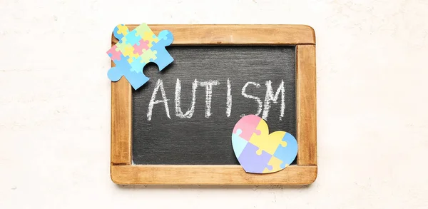 Board with word AUTISM, heart and jigsaw puzzle piece on light background