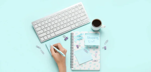 Female hand with pen, notebook, stationery, computer keyboard and cup of coffee on light blue background