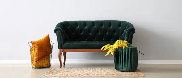 Green sofa, pouf with mimosa flowers and basket with pillows near light wall in room