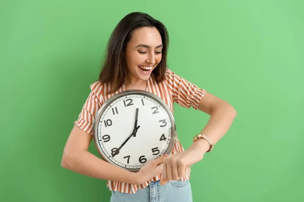 Young woman with wall clock looking at wristwatch on green background