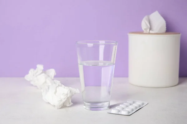 Glass of water with pills and tissue box on table near lilac wall. Allergy concept