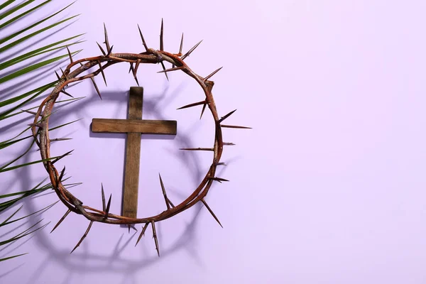 Wooden cross with crown of thorns and palm leaf on lilac background. Good Friday concept