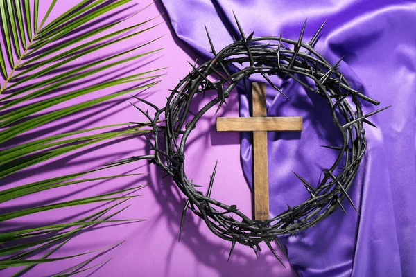 Wooden cross with crown of thorns, palm leaf and purple fabric on violet background. Good Friday concept