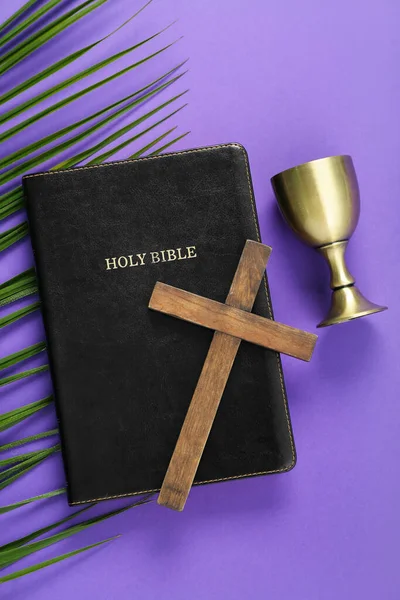 Holy Bible with wooden cross, palm leaf and wine cup on purple background. Good Friday concept