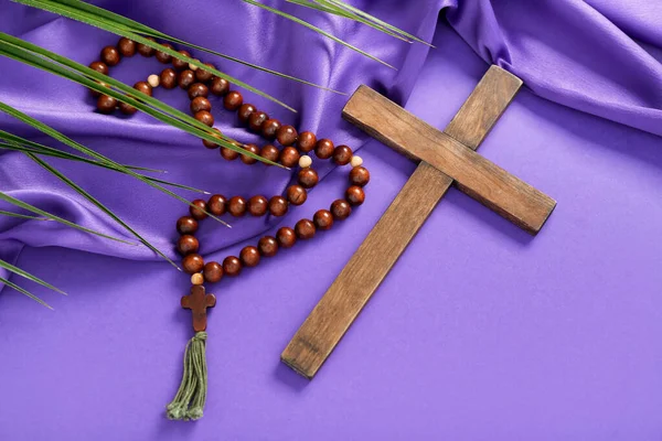 Wooden cross with prayer beads, palm leaf and purple fabric on color background. Good Friday concept