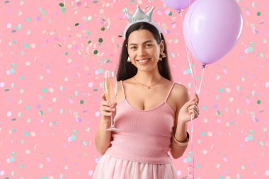 Bir bardak şampanya ve balonla mutlu genç bir kadın pembe arka planda doğum gününü kutluyor.