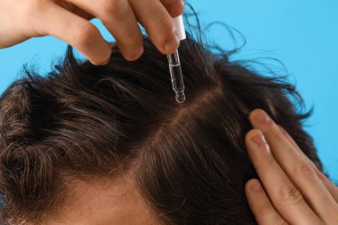Genç adam mavi arka planda saç büyütmek için serum kullanıyor, yakın plan.