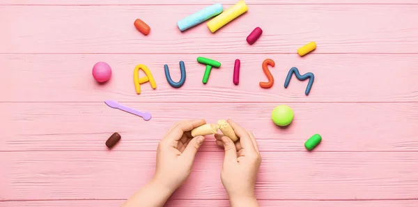 Child\'s hands with plasticine and word AUTISM on pink wooden background