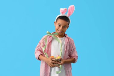 Mavi arka planda Paskalya yumurtası sepeti olan tavşan kulaklı küçük Afro-Amerikalı çocuk.
