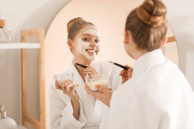 Banyoda aynaya yakın fırçayla zerdeçal maskesi takan genç bir kadın.