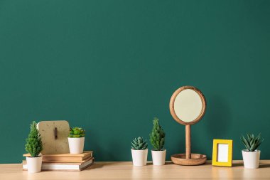 Yeşil duvarın yanındaki masa üzerinde kitap, saat ve ayna bulunan yapay bitkiler.