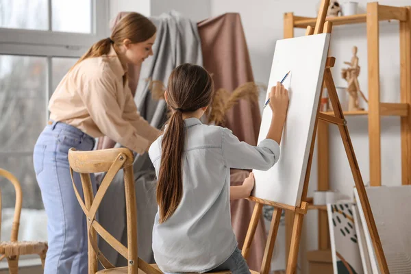 Little girl learning to draw with teacher near easel in workshop