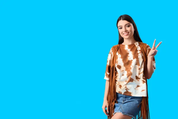 Stylish young woman in tie-dye t-shirt showing victory gesture on light blue background