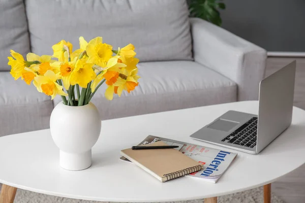 Modern laptop, vase with narcissus flowers, notebook and magazine on coffee table in living room