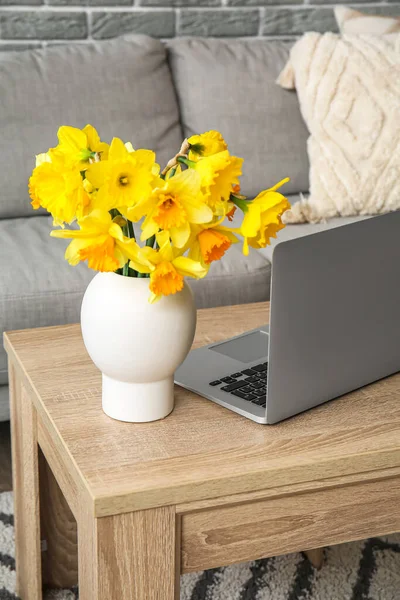 Modern laptop and vase with narcissus flowers on coffee table in living room