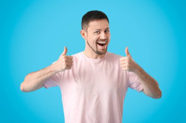 Mavi arka planda baş parmak gösteren tişörtlü yakışıklı bir adam.