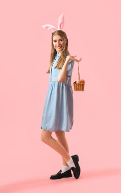 Pembe arka planda Paskalya yumurtası sepeti olan tavşan kulaklı genç bir kadın.