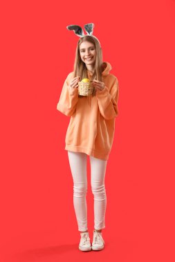 Kırmızı arka planda Paskalya yumurtası sepeti olan tavşan kulaklı genç bir kadın.