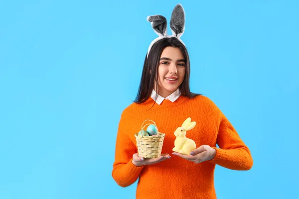 Young woman in bunny ears with basket of Easter eggs and toy on blue background