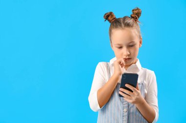 Cep telefonlu küçük bir kız mavi arka planda tırnaklarını yiyor.