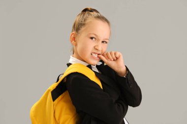 Little schoolgirl with backpack biting nails on grey background