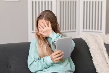 Evdeki koltukta tablet bilgisayarı olan korkmuş küçük bir kız.