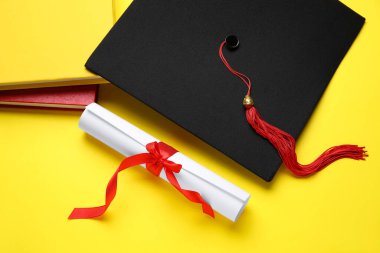 Kırmızı kurdeleli diploma, mezuniyet şapkası ve sarı arka plan kitapları.