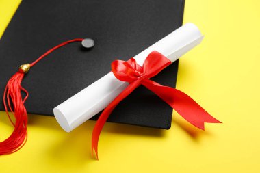 Kırmızı kurdeleli diploma ve sarı arka planda mezuniyet şapkası.