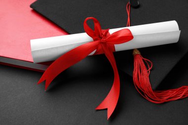 Kırmızı kurdeleli diploma, mezuniyet şapkası ve siyah arka plan kitabı.